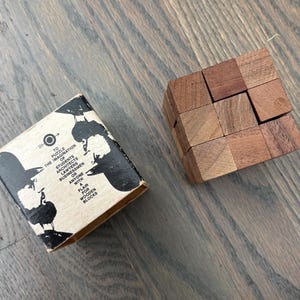 May include: A wooden block puzzle consisting of nine interconnected cubes with varying brown wood grain patterns, accompanied by a square cardboard box featuring black graphic illustrations and the text: TO PUZZLE THE IMAGINATION OF STUDENTS ARCHITECTS LAWYERS BUSINESSMEN OR ANYONE WITH A FLAIR FOR WOODEN BLOCKS.