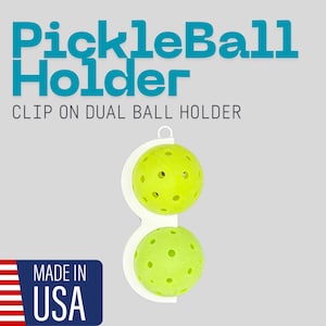 Puede incluir: Soporte de bolas doble blanco con clip con dos pelotas de pickleball verde neón. El texto "PickleBall Holder" está en turquesa encima del soporte. El texto "CLIP ON DUAL BALL HOLDER" está debajo. Bandera americana y "MADE IN USA" abajo a la izquierda.