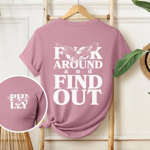 May include: A pink t-shirt with a white eagle graphic and the text "F**k Around and Find Out".