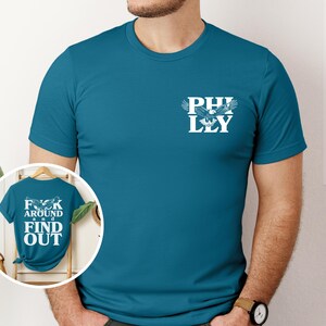 May include: A teal blue t-shirt with a white eagle graphic and the text "PHILLY" on the front. The back of the shirt has the text "F**K AROUND AND FIND OUT".