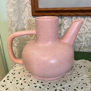 May include: A pink ceramic teapot with a speckled glaze and a wide spout. The teapot has a rounded body and a handle on the side.