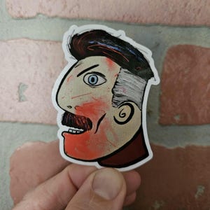 May include: A sticker of a cartoon man's face with a red and white painted look. The man has a mustache and a blue eye. The sticker is on a brick background.