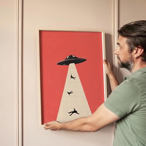 Puede incluir: Impresi&oacute;n art&iacute;stica enmarcada con una ilustraci&oacute;n retro de un OVNI abduciendo figuras. La obra de arte tiene un fondo rojo, un OVNI negro y figuras en un haz de luz blanco. El marco es de color madera clara.