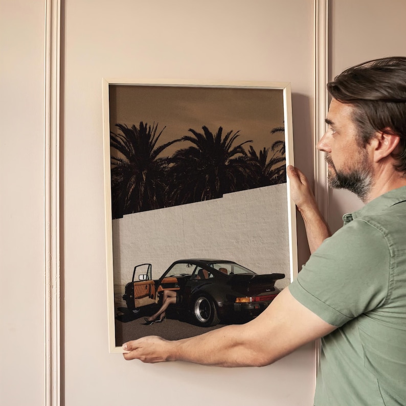 A moody vintage Porsche sports car with an open door and a subtle human presence is set against a white wall and silhouetted palm trees in this cinematic retro art print. The print features muted warm tones and deep shadows for a nostalgic atmosphere. Displayed in a contemporary living space, it adds classic car wall art with elegant, vintage appeal.