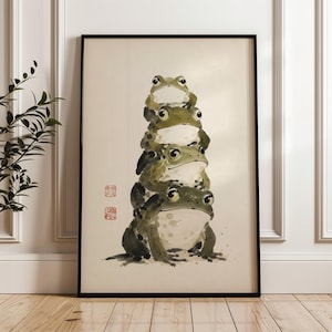 May include: A framed vertical art print featuring a watercolor illustration of four green frogs stacked vertically on top of each other. The frogs have light-colored bellies and dark green backs. Two red square stamps are visible on the left side.