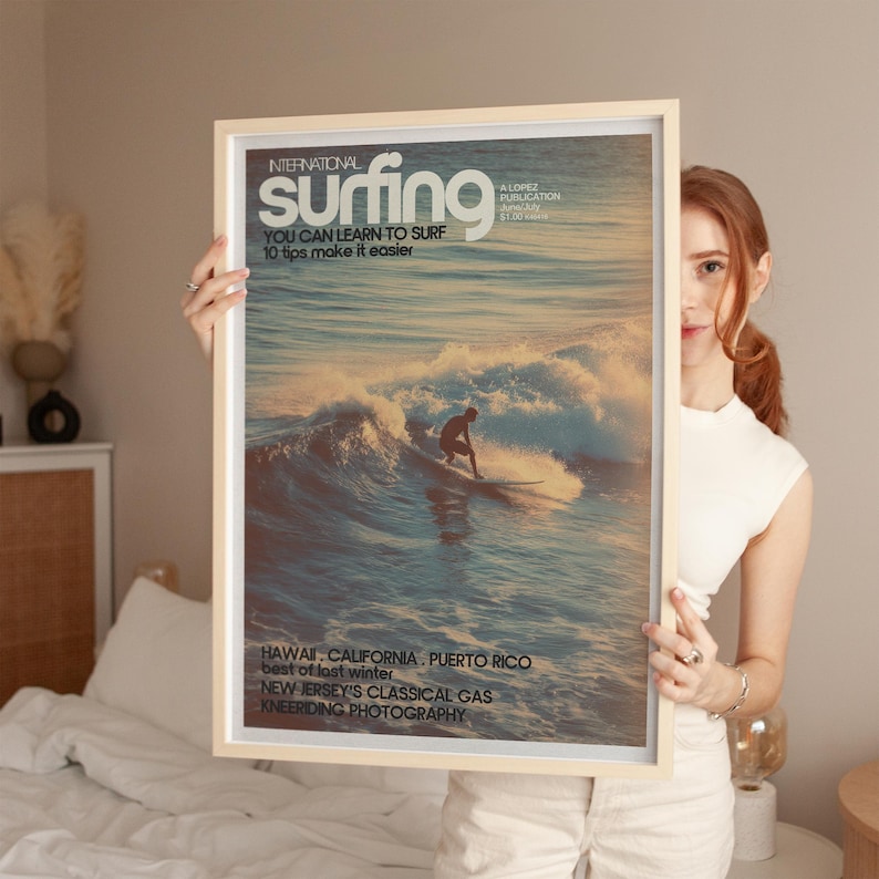 A vintage coastal surf poster featuring a lone surfer riding a wave bathed in golden light with retro typography. The artwork evokes a classic, nostalgic beach vibe with soft pastel ocean hues. Displayed in a warm, cozy bedroom setting.