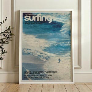 A vintage surf poster featuring a lone surfer riding a large ocean wave in cool blues and whites, framed and displayed above a desk in a sunlit room. The retro style evokes a nostalgic coastal vibe perfect for beach-themed decor. Soft natural light highlights the calming energy of the surfing scene.