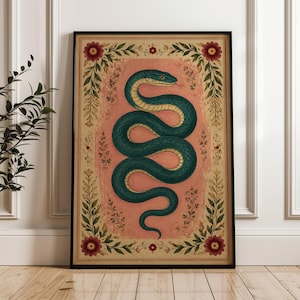 May include: A framed rectangular wall art print featuring a central coiled teal snake on a textured dusty pink background. The artwork is enclosed by a decorative border of green leafy vines and small red flowers on a cream-coloured parchment base.