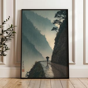 May include: Framed art print depicting a rainy mountain scene. A lone figure with an umbrella walks along a path, with mountains in the background. The artwork is in a traditional Japanese style, with muted colors and a sense of tranquility.