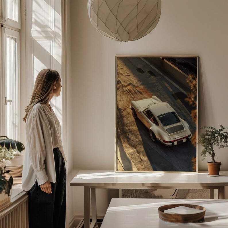 A vintage Porsche 911 captured in warm golden-hour light, highlighting the car's sleek curves and classic design. The automotive wall art print features nostalgic shadows and earthy tones, perfect for car enthusiasts. Displayed on a table in a bright, minimalist living space.