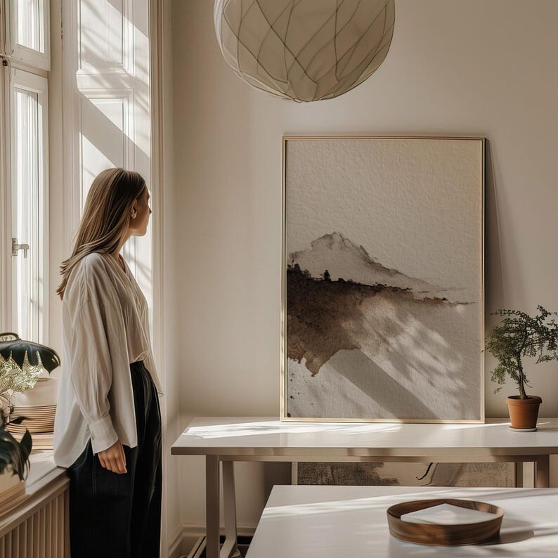 A minimal abstract landscape in earthy brown and beige watercolor tones with a wabi-sabi aesthetic. The serene, textured art print is displayed in a bright, modern room filled with natural light. It creates a calm and rustic atmosphere, ideal for minimalist or zen-inspired interiors.