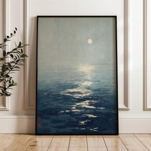 May include: A framed vertical wall art print depicting a serene seascape at night. The scene features a pale, circular moon suspended in a muted gray sky above a dark blue ocean, with a bright, shimmering reflection of moonlight on the water surface.