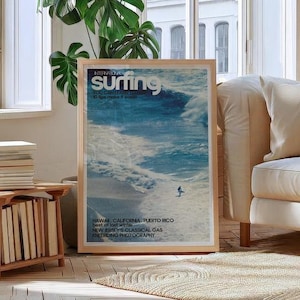 A vintage surf poster featuring a lone surfer riding expansive ocean waves in vibrant blue tones with retro magazine-style text. The coastal wall art print evokes a nostalgic beach vibe and sense of adventure. Displayed in a bright, cozy living room setting.