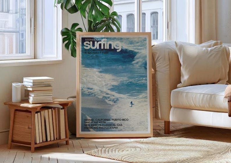 A vintage surf poster featuring a lone surfer riding expansive ocean waves in vibrant blue tones with retro magazine-style text. The coastal wall art print evokes a nostalgic beach vibe and sense of adventure. Displayed in a bright, cozy living room setting.