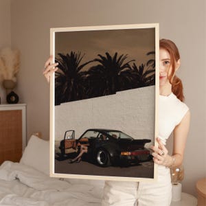 A vintage black Porsche sports car with an open door and a figure’s legs visible, set against a white wall and silhouetted palm trees in moody, warm tones. The cinematic retro poster captures a sleek, nostalgic vibe. Displayed in a cozy bedroom environment.