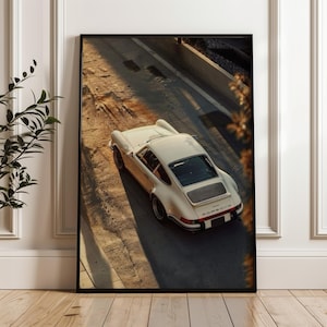 A vintage Porsche 911 captured in warm, golden-hour light highlighting its sleek curves and classic design. This automotive wall art print features nostalgic earthy tones and soft shadows. Displayed leaning against a wall in a modern living room setting.