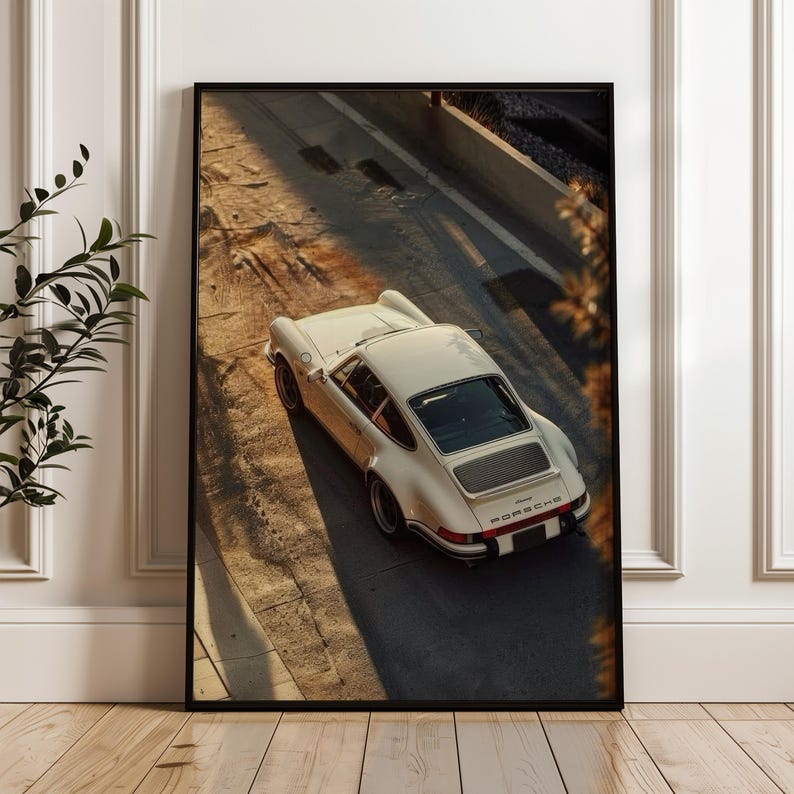 A vintage Porsche 911 captured in warm, golden-hour light highlighting its sleek curves and classic design. This automotive wall art print features nostalgic earthy tones and soft shadows. Displayed leaning against a wall in a modern living room setting.