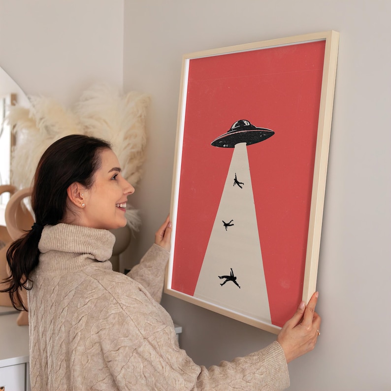 Puede incluir: Impresi&oacute;n art&iacute;stica enmarcada con una ilustraci&oacute;n retro de un OVNI negro que absorbe tres figuras silueteadas sobre un fondo rojo. La impresi&oacute;n est&aacute; en un marco de madera clara, perfecta para a&ntilde;adir un toque de fantas&iacute;a a cualquier espacio.