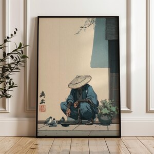 May include: Framed art print featuring an illustration of a person in traditional Japanese attire, squatting near a bowl and pigeons. The person wears a straw hat and blue clothing. The background is a beige wall with a blue accent and a wooden door.