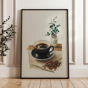 May include: Watercolor print of a black coffee cup and saucer, with coffee beans on a spoon. A small glass jar with green sprigs sits next to the cup. The artwork is framed in a black frame.