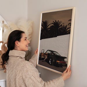 A moody vintage Porsche sports car with an open door is captured in a cinematic wall art print featuring muted warm tones and silhouetted palm trees. The retro poster exudes elegance and mystery against a stark white wall. Displayed in a cozy living space, this classic car print adds sophisticated vintage charm.