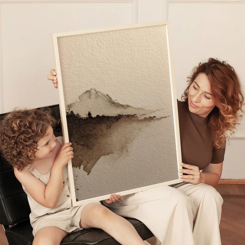 Minimalist abstract landscape art in earthy brown and beige tones with a wabi-sabi style, featuring a subtle watercolor wash on textured paper. The serene and calming fine art print is held by a woman and child in a cozy living space. This poster complements minimalist and rustic interiors with its natural, understated aesthetic.