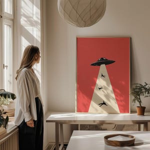 Puede incluir: Una impresi&oacute;n art&iacute;stica enmarcada con un fondo rojo, que representa un OVNI negro y figuras que son levantadas por un rayo de luz. La obra de arte tiene un estilo retro y minimalista, con un marco dorado. La impresi&oacute;n est&aacute; sobre una mesa blanca.