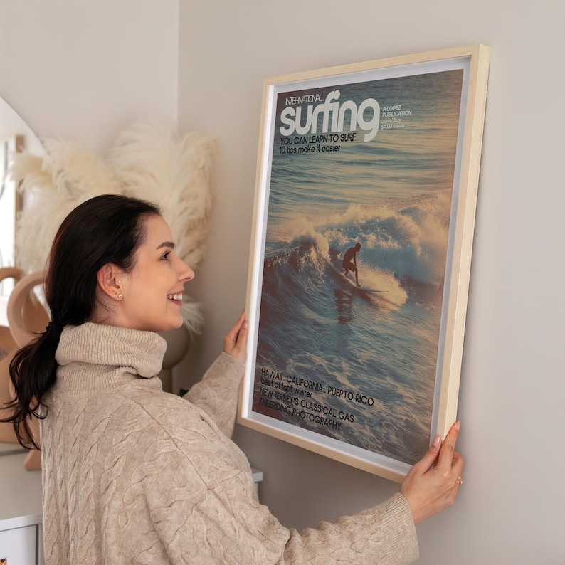 A vintage-style surf poster featuring a lone surfer riding a glowing ocean wave in soft pastel hues. The nostalgic coastal art print has a retro magazine cover design with timeless typography. It hangs on a neutral wall, adding a calming beach vibe to the living space.