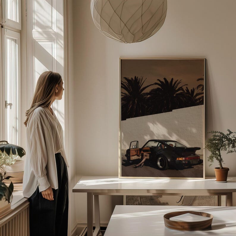 A cinematic wall art print featuring a vintage black Porsche sports car with an open door against a white wall and silhouetted palm trees in warm, moody tones. The retro poster evokes mystery and elegance with deep shadows and muted colors. Displayed above a simple table in a softly lit modern living space.