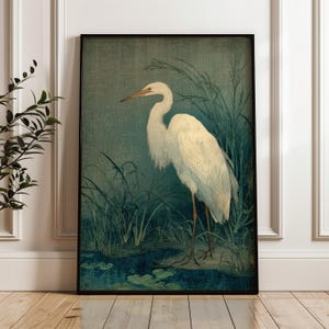 May include: A framed print of a white egret standing in a marsh. The artwork features the bird with a long neck and beak, surrounded by tall green reeds and water lilies. The background is a muted teal color.