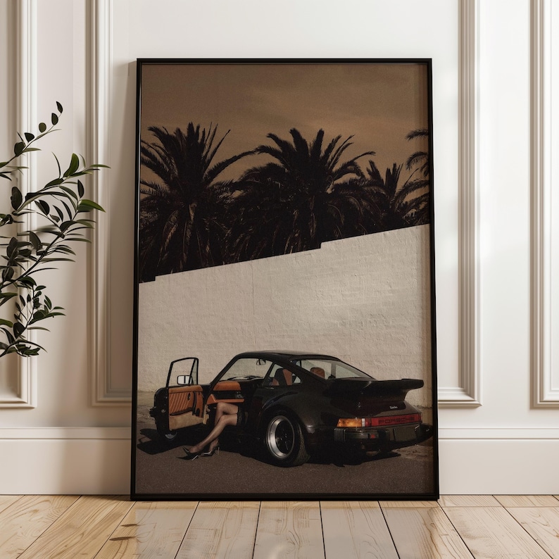 A moody vintage Porsche sports car with an open door and a pair of legs visible inside, set against a stark white wall and silhouetted palm trees in muted warm tones. This cinematic retro wall art print exudes elegance and mystery. Displayed leaning against a white paneled wall in a stylish living space.