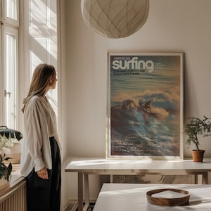 A vintage surf poster showcasing a lone surfer riding a wave with warm, golden light and soft pastel ocean colors. The retro coastal wall art has classic typography and a nostalgic magazine cover style. Displayed above a minimalist desk in a sunlit room, it adds a calming, adventurous vibe.