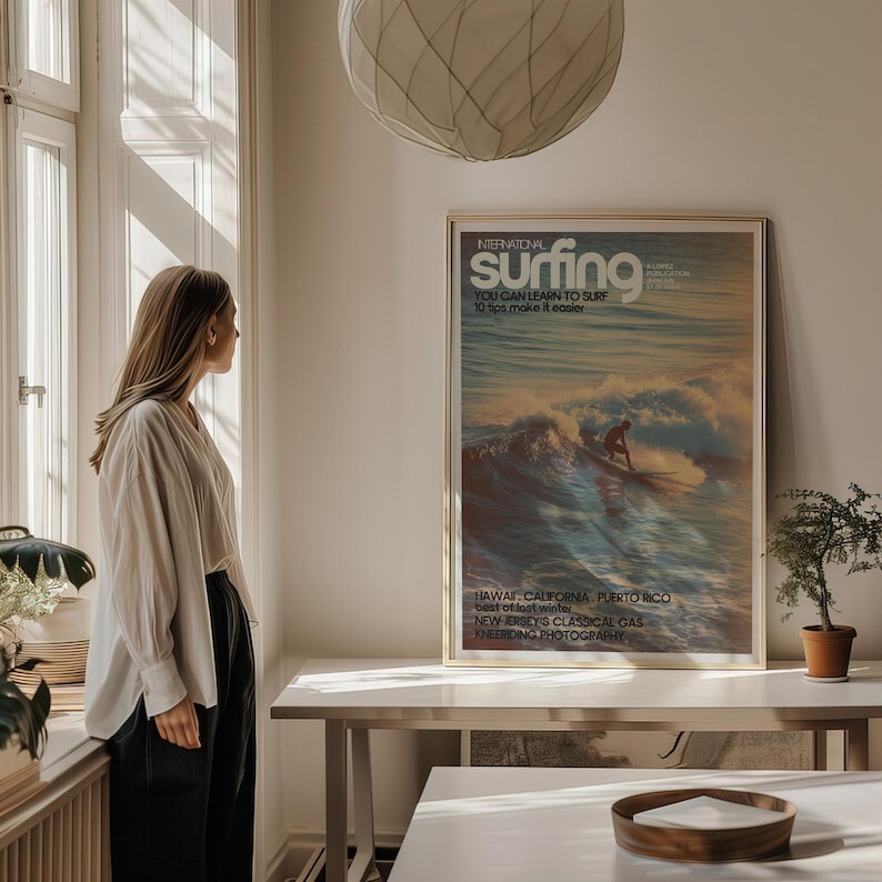 A vintage surf poster showcasing a lone surfer riding a wave with warm, golden light and soft pastel ocean colors. The retro coastal wall art has classic typography and a nostalgic magazine cover style. Displayed above a minimalist desk in a sunlit room, it adds a calming, adventurous vibe.