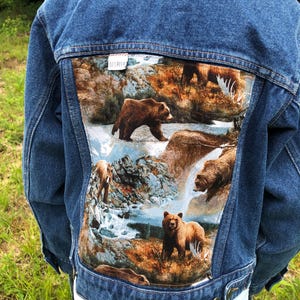 May include: Blue denim jacket with a large, colorful bear scene on the back. The artwork depicts brown bears near a waterfall and river. The jacket has a classic design with button closures and pockets, perfect for casual wear.