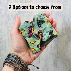 Può includere: Una piccola custodia in tessuto piegato con uno sfondo verde chiaro caratterizzato da un motivo di falene colorate, lune crescenti gialle e foglie verdi. La custodia è chiusa da un bottone a pressione bianco. Il testo 9 Options to choose from è visibile.