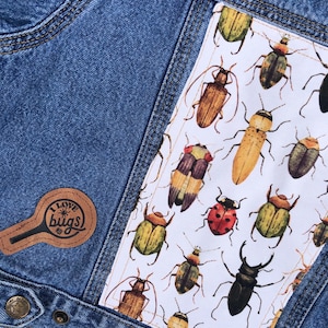 May include: Close-up of a denim jacket with a white patch featuring colorful insect illustrations. A brown leather patch with the text "I LOVE bugs" is on the front. The jacket has a classic design with button closures and pockets.