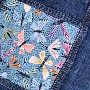May include: Close-up of a denim pocket with a colorful butterfly pattern. The butterflies are in shades of pink, orange, green, and purple, set against a light blue background. The pocket is sewn onto a blue denim fabric.