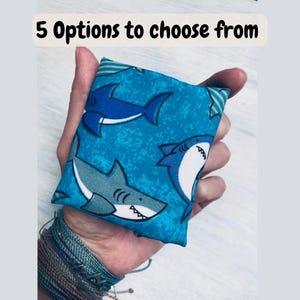 May include: A square fabric pouch featuring a repeating pattern of blue and white sharks on a bright teal background. The text 5 Options to choose from is displayed at the top of the image. The pouch is held in a palm, showing its compact size.