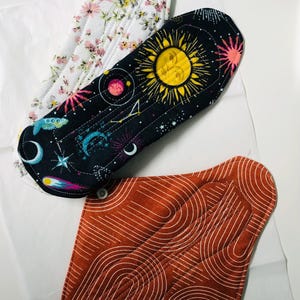 May include: Three reusable cloth menstrual pads. One pad features a white background with a colorful floral pattern. Another pad has a black background with yellow suns, blue moons, and stars. The third pad is rust-colored with white line art.