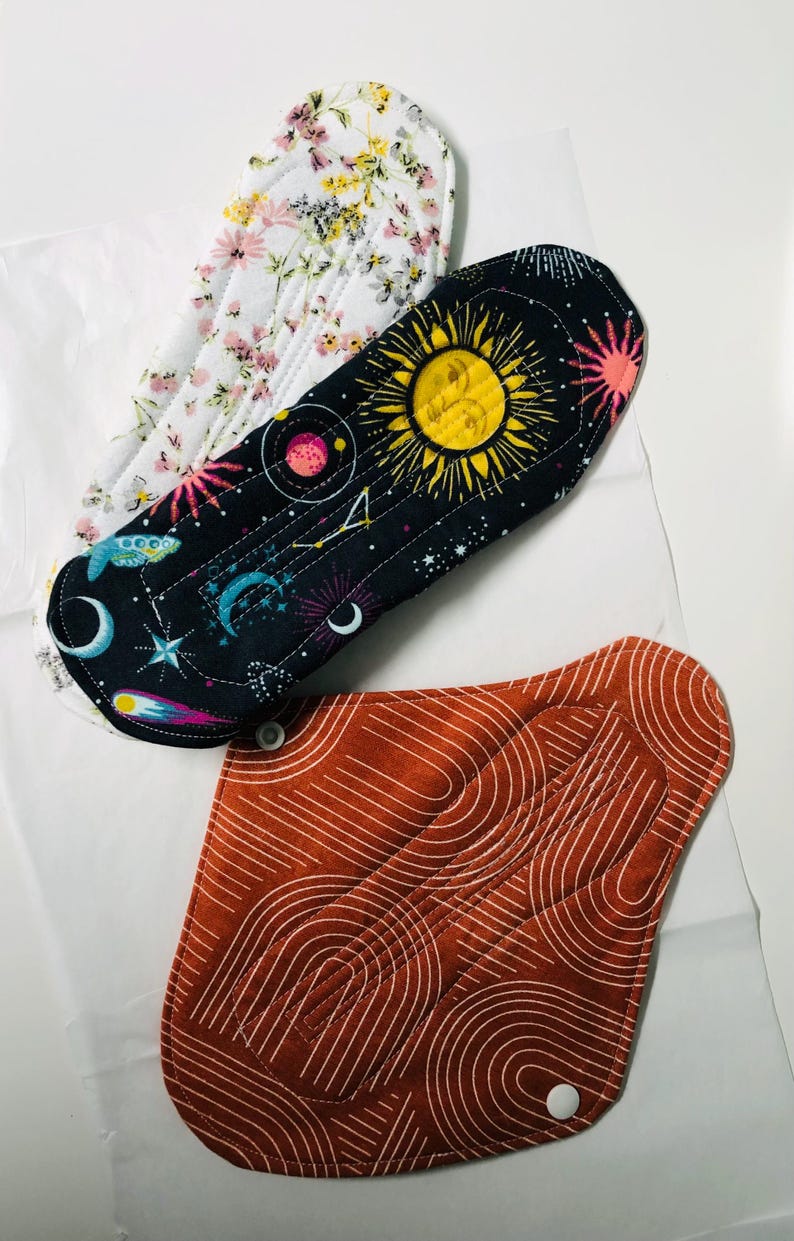 May include: Three reusable cloth menstrual pads. One pad features a white background with a colorful floral pattern. Another pad has a black background with yellow suns, blue moons, and stars. The third pad is rust-colored with white line art.