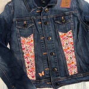 May include: A blue denim jacket with floral fabric panels and a small butterfly accent. The jacket features a patch with the words "pretty, fierce, strong, kind". The jacket has button closures and pockets.