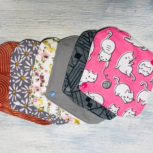 May include: A set of six square fabric pads with rounded corners, featuring various patterns including terracotta arches, grey daisies, floral prints, solid grey, black geometric lines, and a pink background with white cartoon cats and yarn.