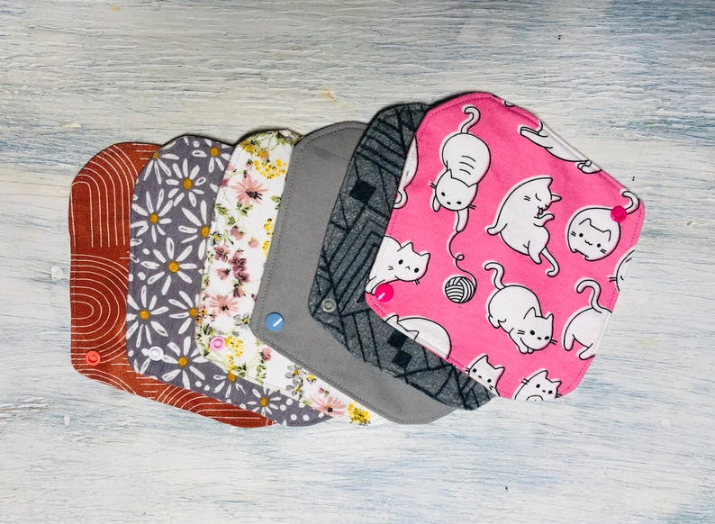 May include: A set of six square fabric pads with rounded corners, featuring various patterns including terracotta arches, grey daisies, floral prints, solid grey, black geometric lines, and a pink background with white cartoon cats and yarn.