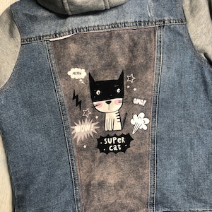 May include: Blue denim jacket with gray hood and sleeves. The back features a gray panel with a cartoon cat graphic, speech bubbles saying "Meow", "Wow", and "OMG!", and the text "Super Cat".