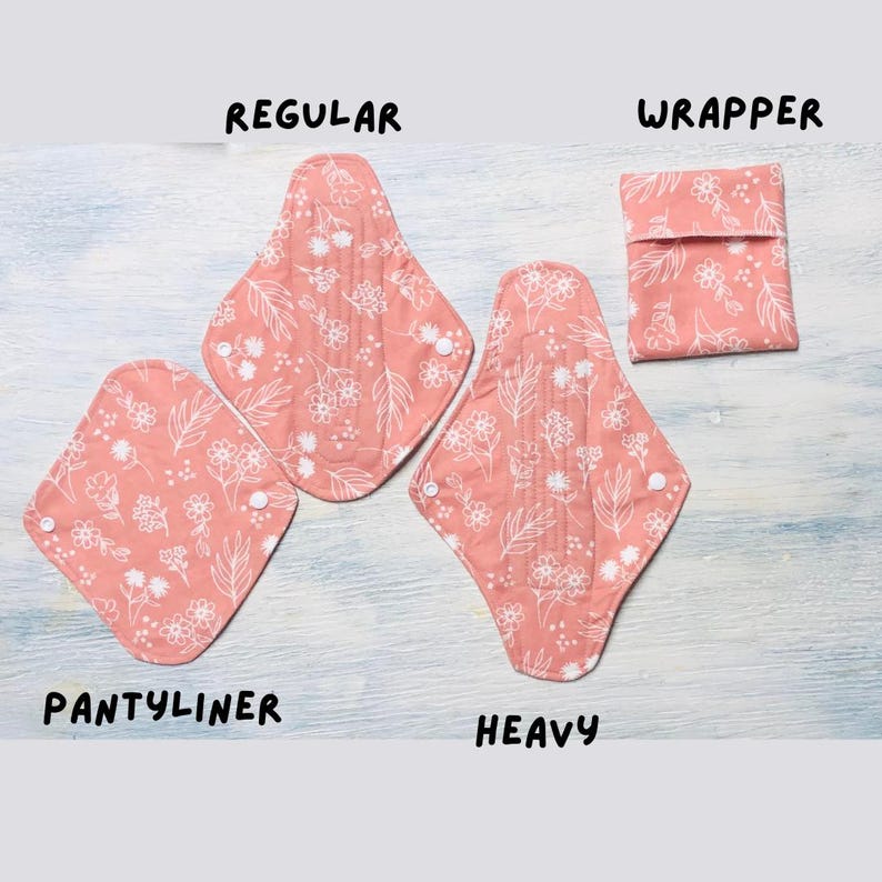 May include: Three reusable cloth menstrual pads and a matching fabric wrapper in a coral pink color with a white floral and leaf line art pattern. The items are labeled as PANTYLINER, REGULAR, HEAVY, and WRAPPER, featuring white snap closures.