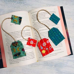Può includere: Cinque segnalibri in tessuto con diversi motivi. I segnalibri sono attaccati a uno spago e pendono da un libro. I motivi includono foglie verdi, banane gialle, fiori rossi e gialli e farfalle verdi.