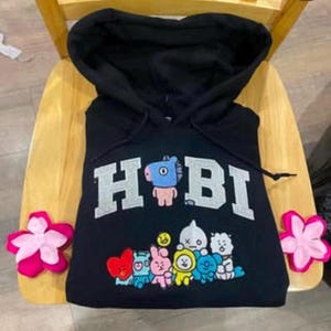 May include: Black hoodie with a white "Hobi" graphic and a group of cartoon characters. The characters are colorful and include a pink bunny, a yellow chick, and a blue bear.