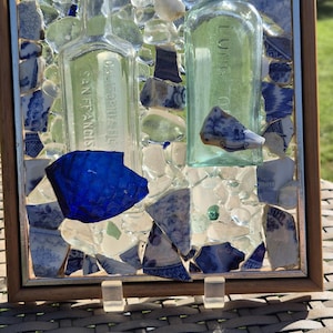 Könnte beinhalten: Ein gerahmtes Mosaikkunstwerk mit zwei antiken Glasflaschen und Fragmenten aus blau-weißem Porzellan. Das Mosaik ist vor einem Hintergrund aus klaren Glasteilen und einem Holzrahmen angeordnet. Die Flaschen sind mit "SAN FRANCISCO" und "LUNG TONIC" beschriftet.