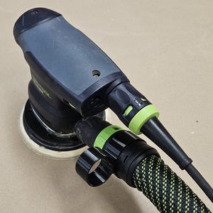 Puede incluir: Una lijadora eléctrica negra y verde con una manguera de aspiración de polvo. La lijadora tiene un gatillo verde y un mango negro.