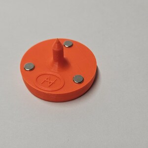 May include: Orange plastic base with a pointed top and three small silver magnets. The base has a circular logo with the letter "H" inside.